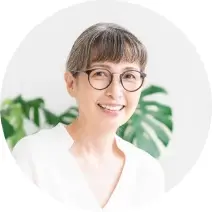 白いVネックの服を着て明るめの色の髪をまとめたメガネをかけたにこやかな50代女性の写真